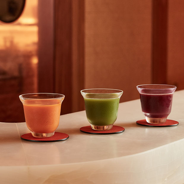 Three small glasses of orange, green and purple wellness juices on coasters atop a light counter