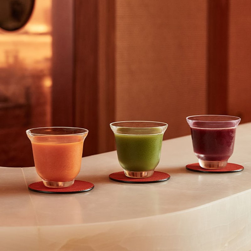 Three small glasses of orange, green and purple wellness juices on coasters atop a light counter