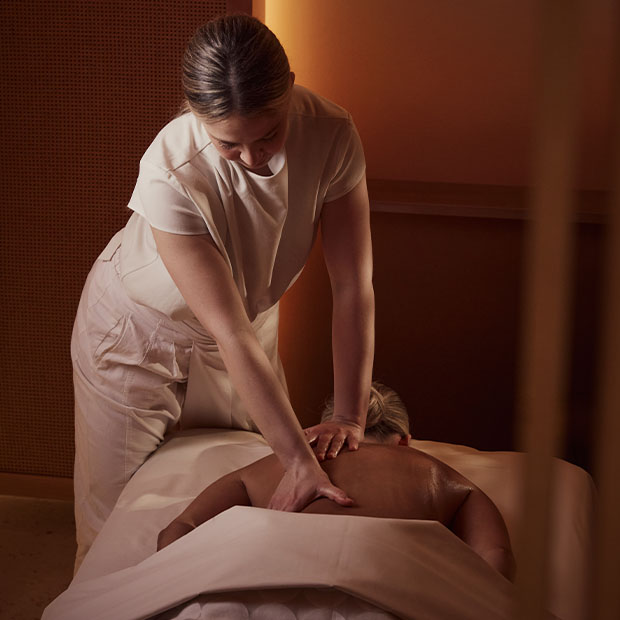 Therapist performing a relaxing back massage on a person lying under a white sheet in a warm spa setting.