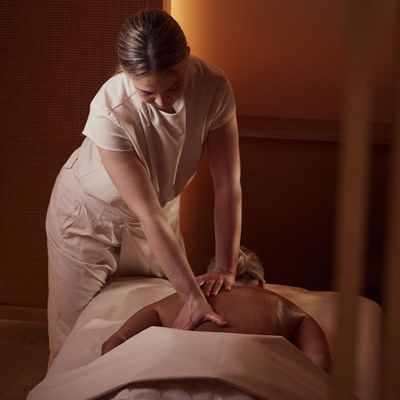 Therapist performing a relaxing back massage on a person lying under a white sheet in a warm spa setting.