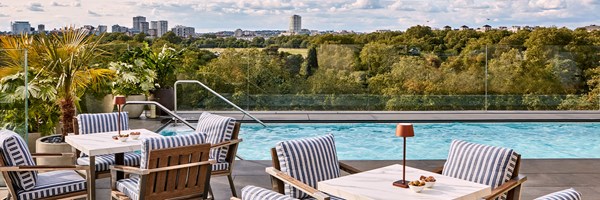The Berkeley rooftop pool, with small square marble tables with blue & white striped chairs tucked in, and a view behind the pool of Hyde Park and tall urban buildings.